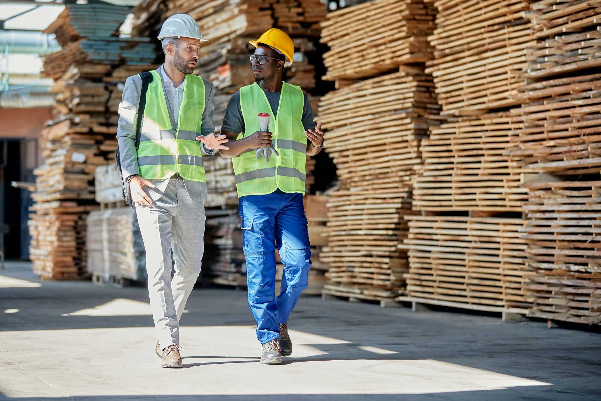 Twee mannen met veiligheidshesjes en helm op in een logistieke omgeving met pallets, samen in gesprek